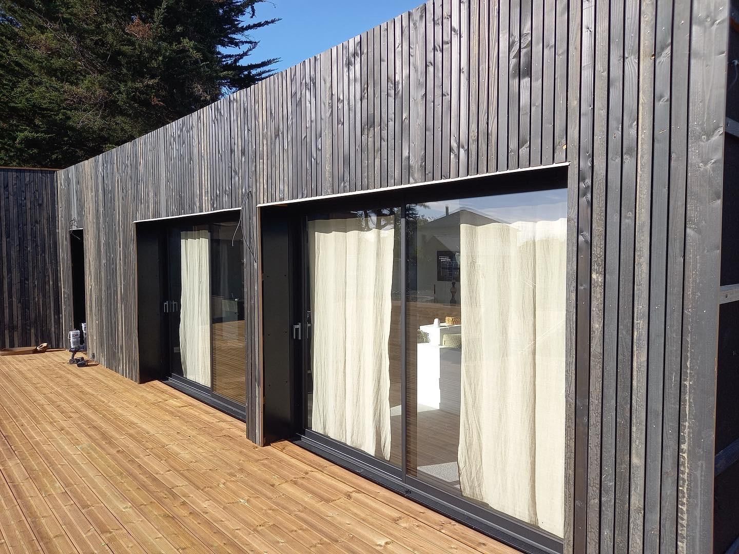 Maison moderne à l'extérieur revêtu de bois, avec de grandes fenêtres et des rideaux, jouxtant une terrasse en bois à claire-voie.