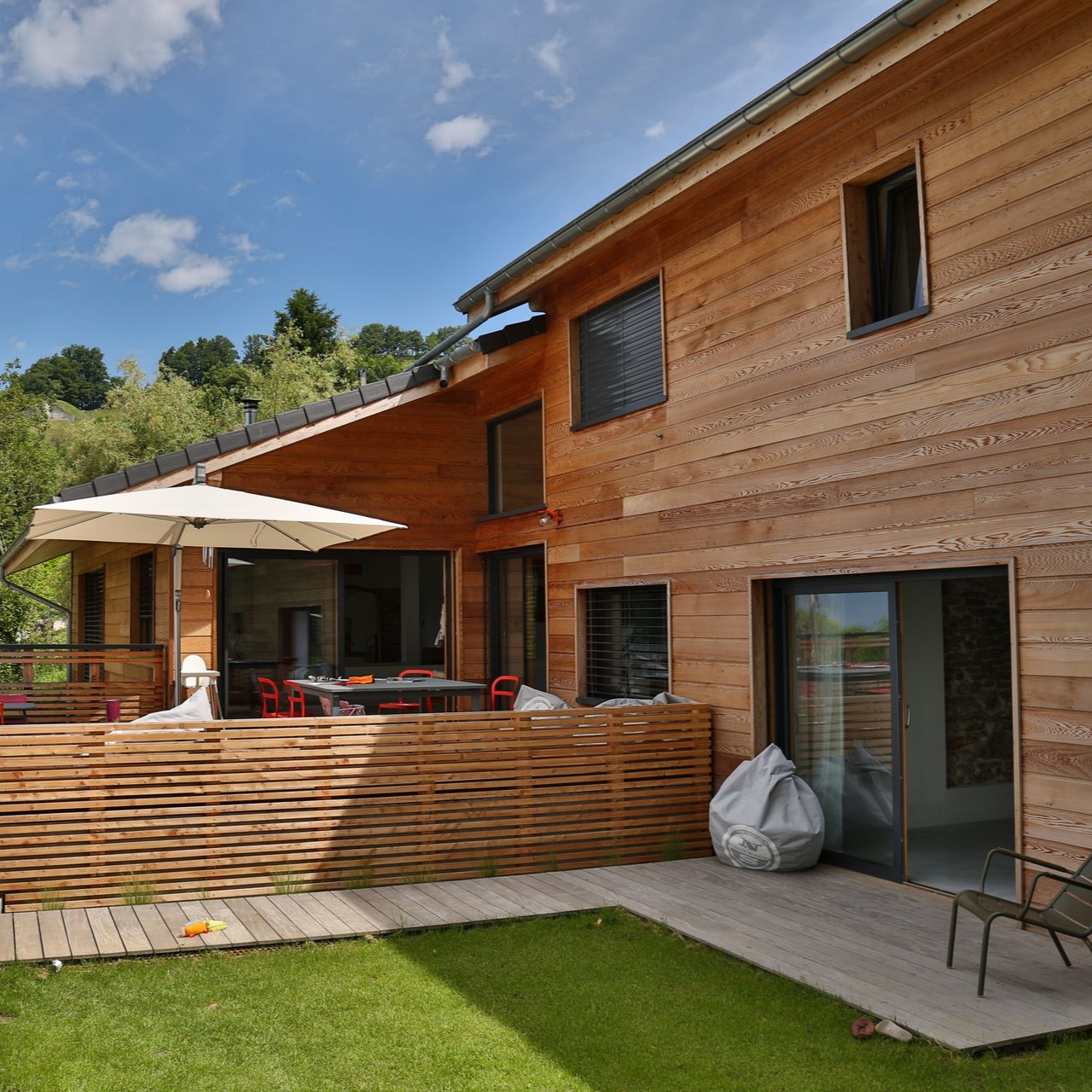 Maison moderne en bois avec patio, portes coulissantes en verre et pelouse verdoyante sous un ciel bleu.