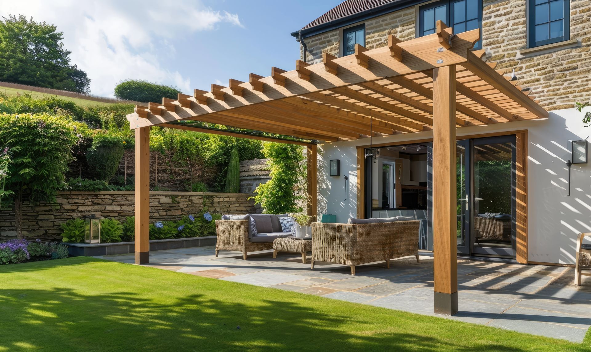 Pergola en bois surplombant une terrasse jouxtant une maison en pierre, avec des sièges extérieurs et une pelouse verdoyante.