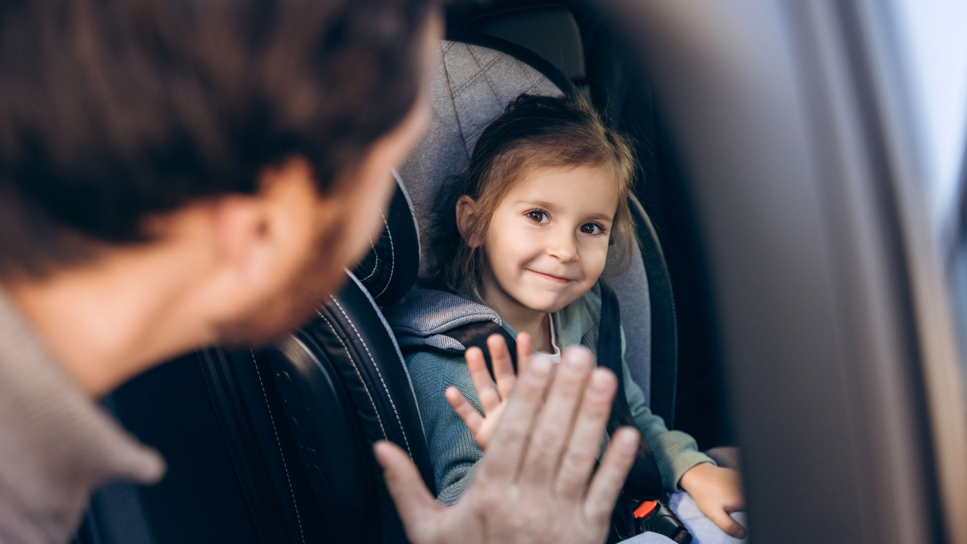 Une personne salue de la main un enfant souriant, installé dans un siège auto à l'intérieur d'un véhicule.