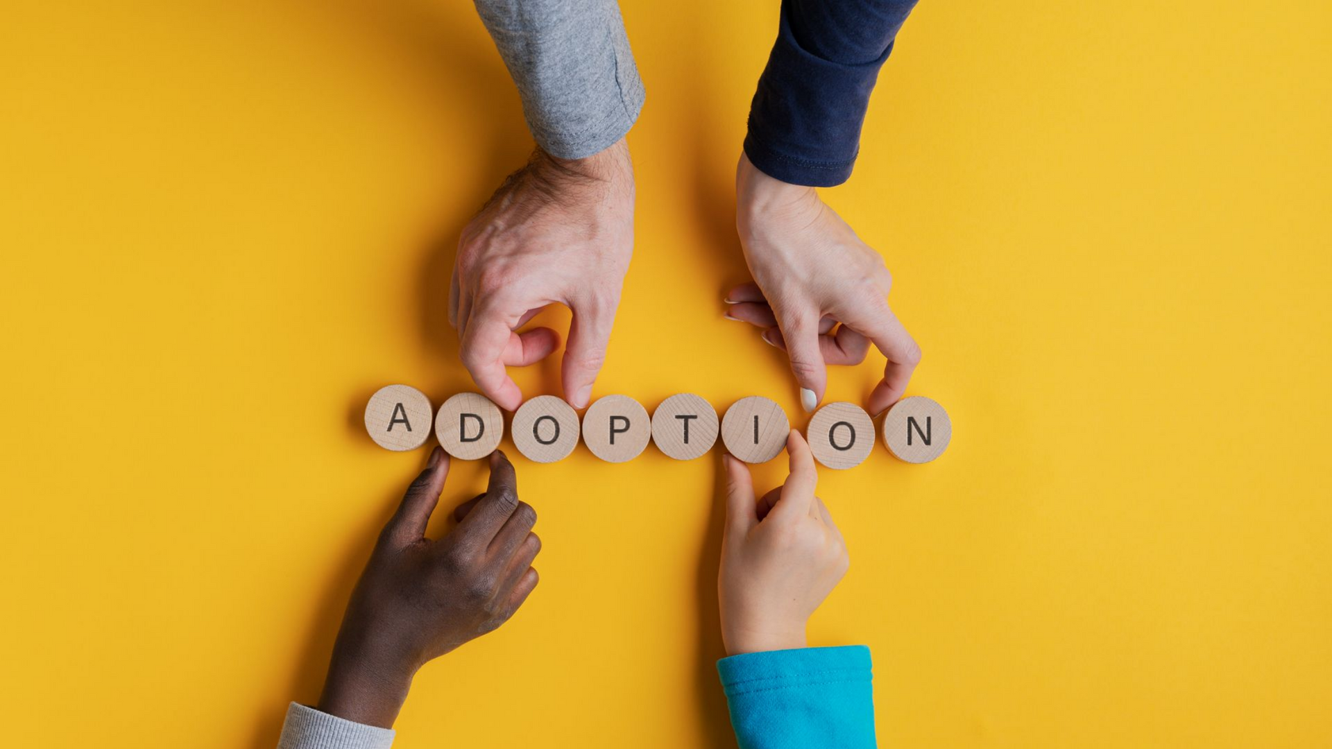 Des mains aux teints variés disposent des lettres en bois formant le mot ADOPTION  sur une surface jaune.