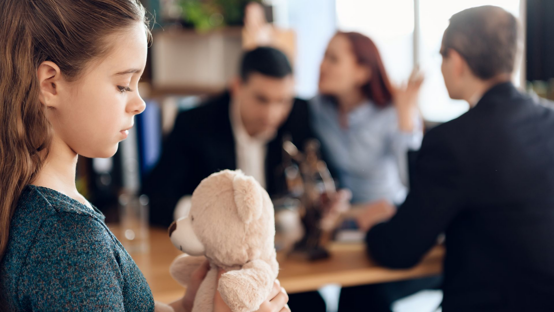 Une fillette avec un ours en peluche, l'air triste, regarde ses parents se disputer à une table, à côté de leur avocat.