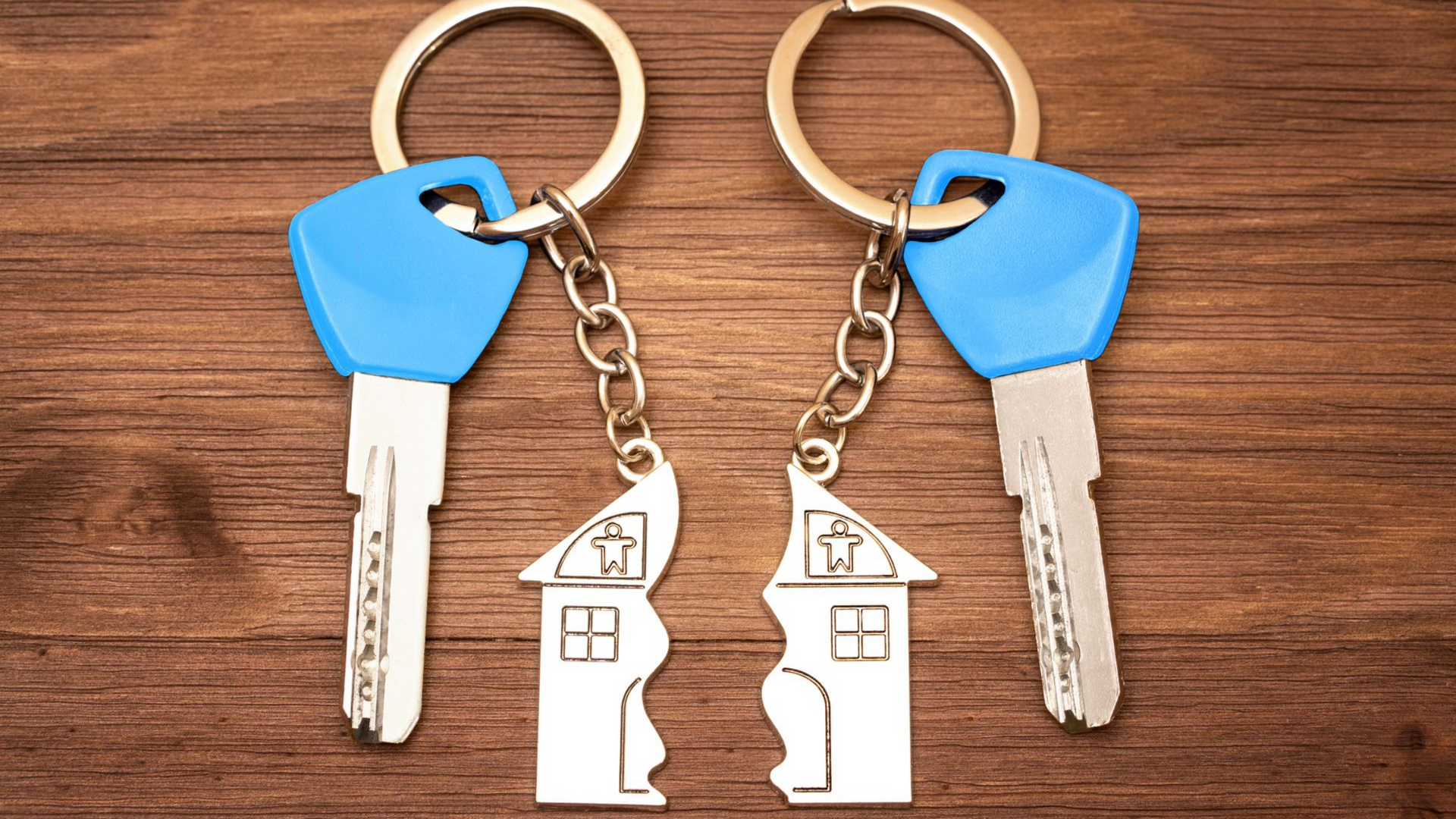 Deux clés à poignées bleues et des porte-clés en forme de maison fendue sur une surface en bois.