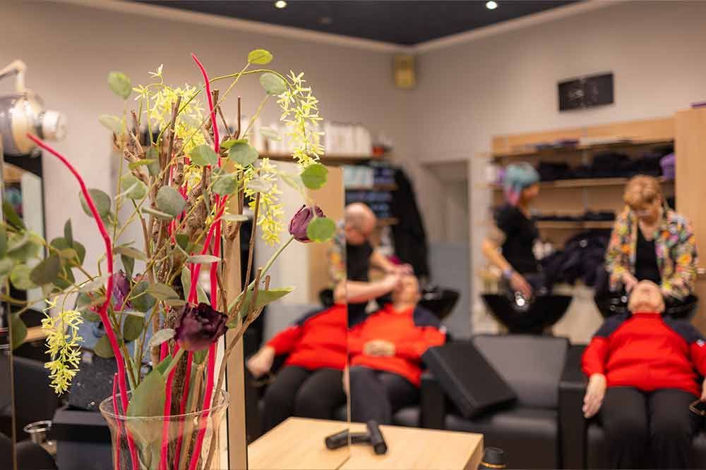 Eine Frau lässt sich in einem Salon die Haare waschen. Im Vordergrund steht eine Vase mit Blumen.