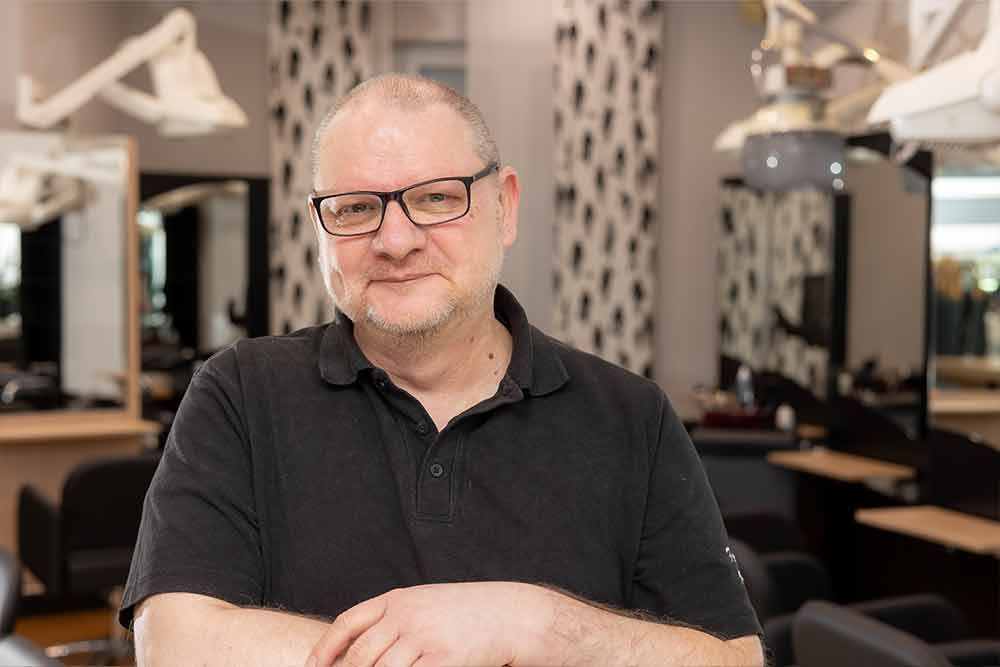 Eine Frau mit Brille und schwarzem Hemd sitzt an einem Tisch in einem Salon.