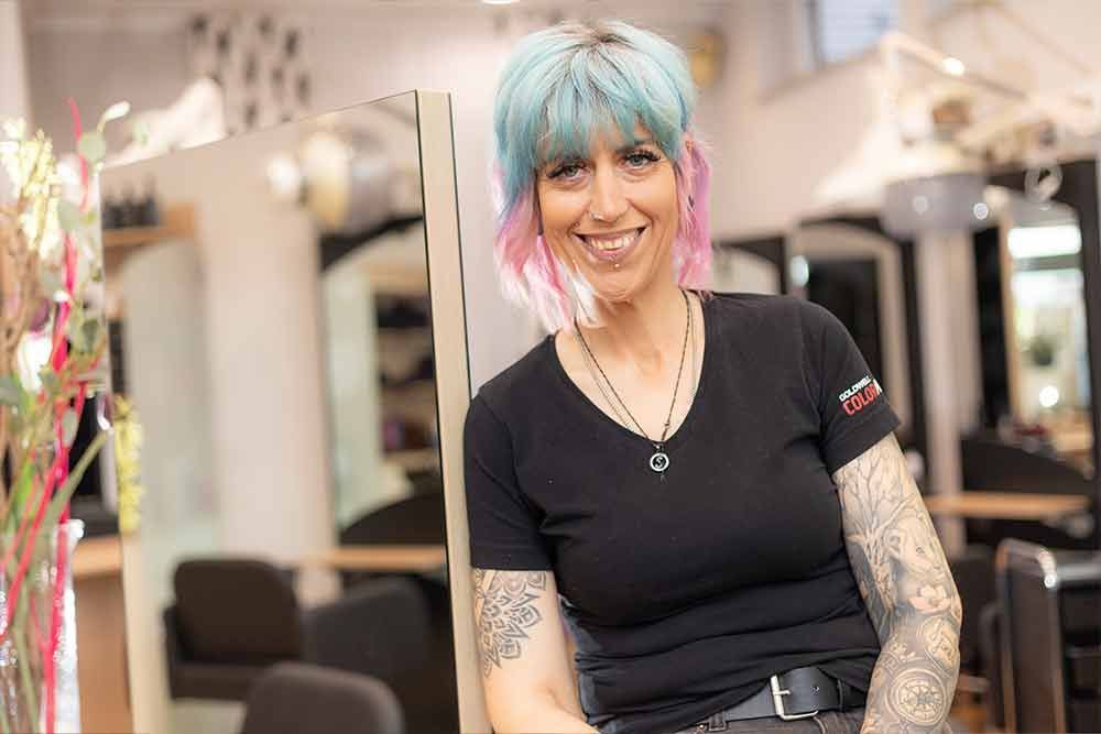 Eine Frau mit blauen und rosa Haaren lächelt in einem Friseursalon.