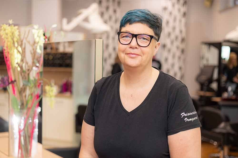 Eine Frau mit Brille und schwarzem Hemd sitzt an einem Tisch in einem Salon.