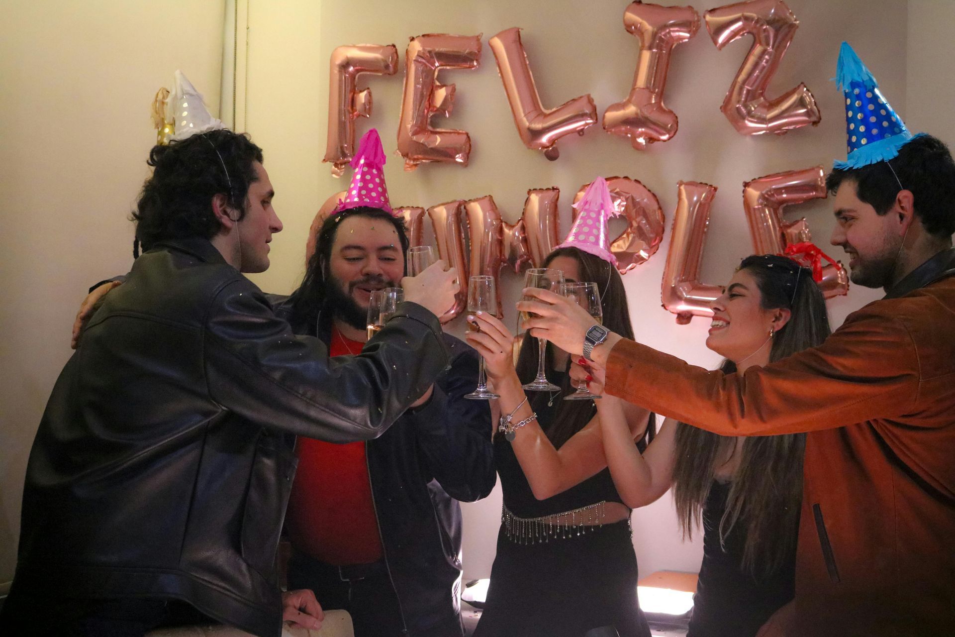 Unos amigos brindan en una fiesta frente a letras de globos color oro rosa y un sombrero de fiesta azul.