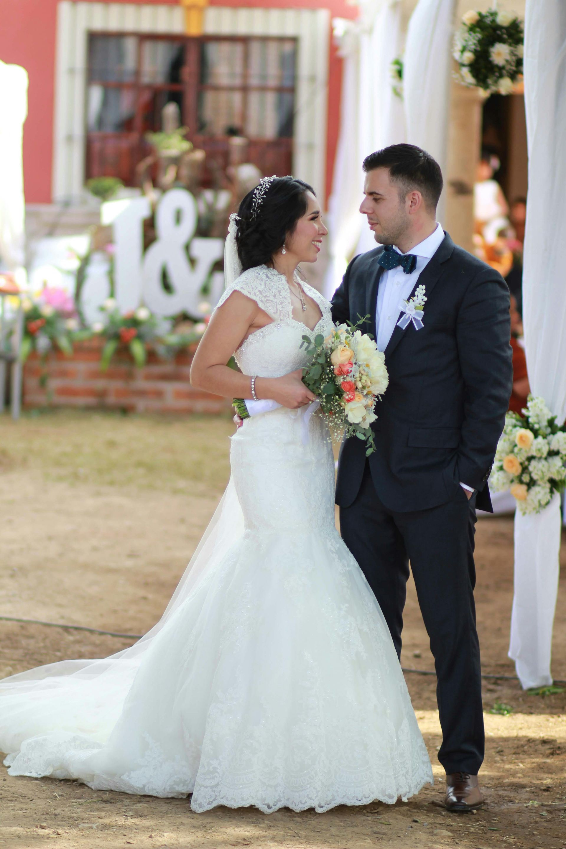 Los novios, de pie al aire libre en una ceremonia nupcial, sonríen y sostienen un ramo.