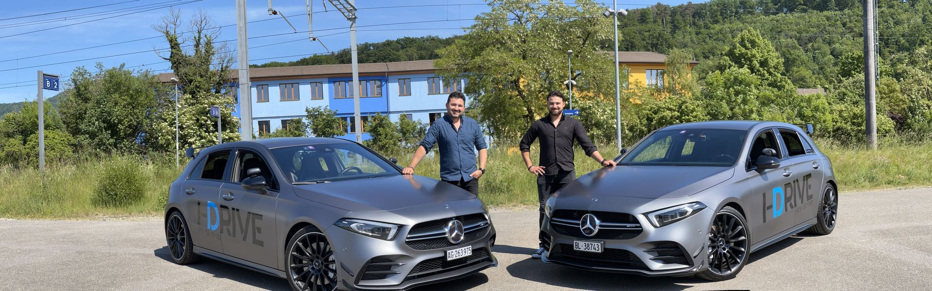 Zwei graue Mercedes-Fahrzeuge parkten im Freien auf einem sonnigen Industriegelände, zwischen ihnen standen zwei Personen.