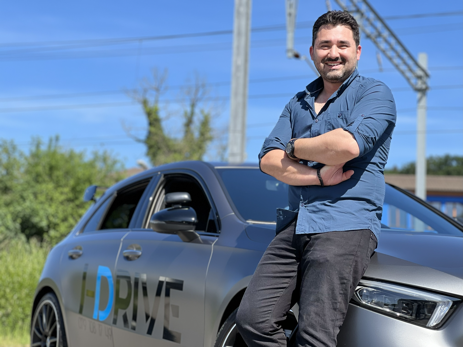Ein lächelnder Mann lehnt an einem grauen Auto mit grosser blauer Aufschrift, im Freien unter klarem blauen Himmel.