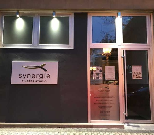 Exterior del estudio Synergie Pilates con ventanas iluminadas y entrada de cristal al anochecer.