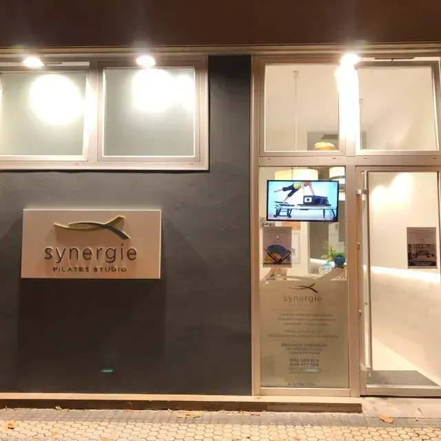 Fachada nocturna del estudio Synergie Pilates con ventanas iluminadas y puerta de cristal.