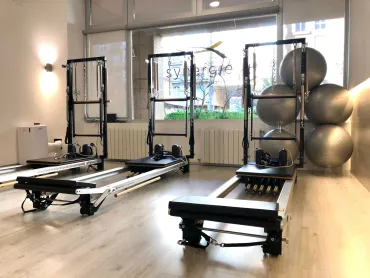 Una mujer se estira en una máquina de Pilates en un gimnasio luminoso, con otra persona haciendo ejercicio al fondo.
