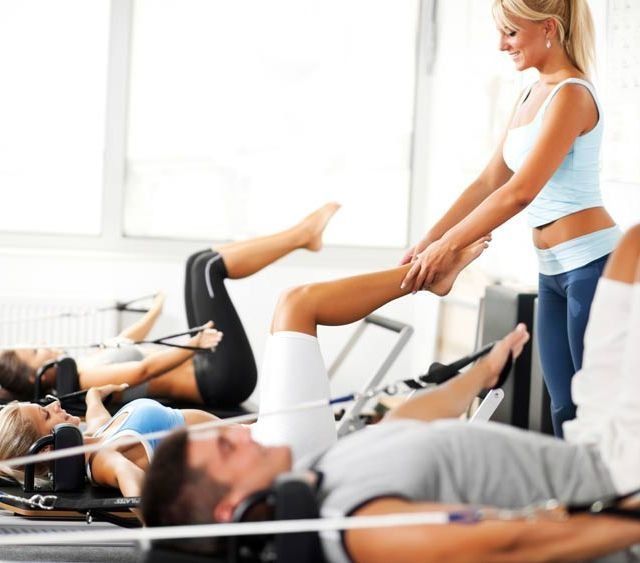 Instructor de fitness estirando las piernas de un cliente durante una sesión de entrenamiento grupal en camas reformer.