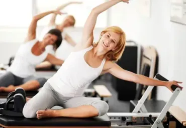 Una mujer se estira en una máquina de Pilates en un gimnasio luminoso, con otra persona haciendo ejercicio al fondo.