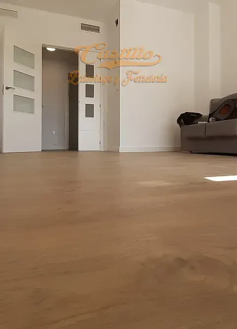 Habitación vacía con suelo de madera, paredes blancas y dos puertas. Un sofá gris está en la esquina.