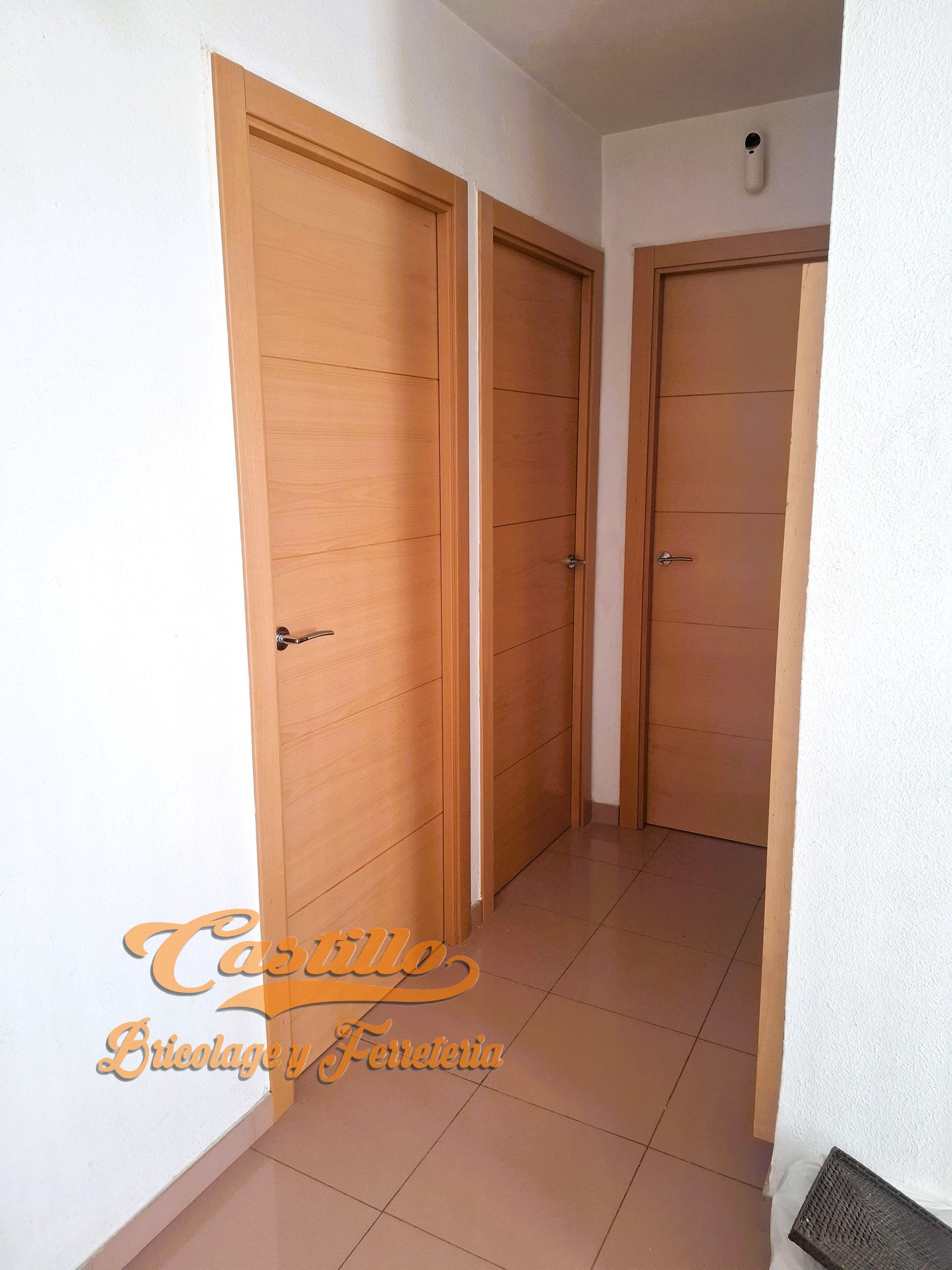 Un pasillo con tres puertas de madera cerradas. Las paredes son blancas y el suelo es de baldosas.