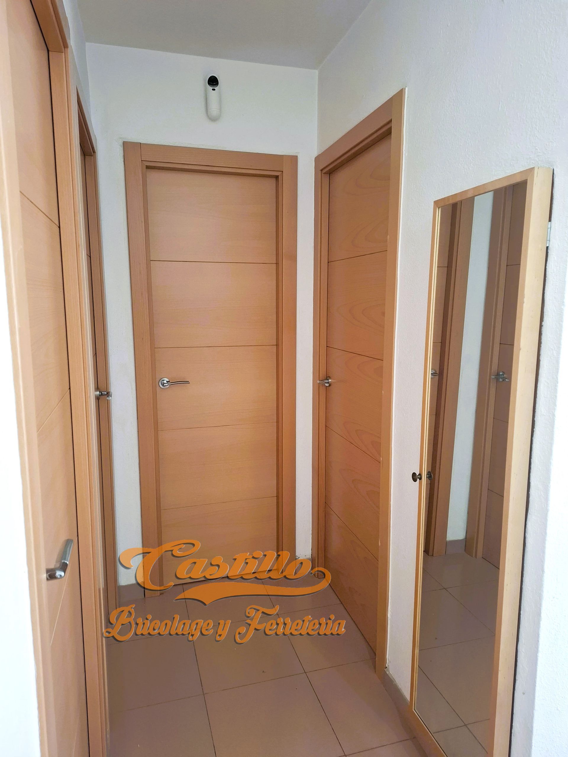 Pasillo estrecho con puertas de madera clara, un espejo y una pared blanca.