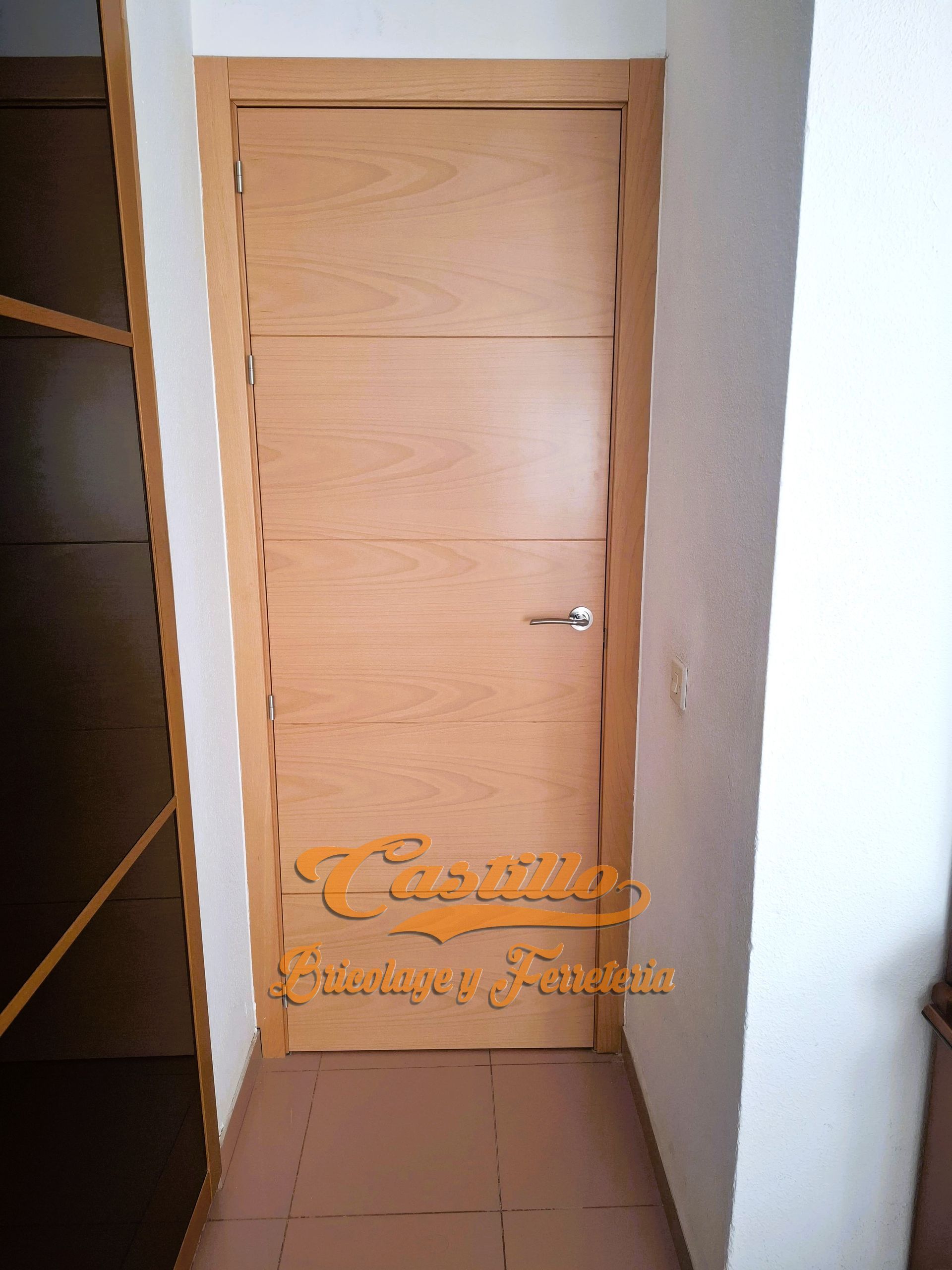Una puerta de madera clara en un pasillo, con tirador plateado y paredes de color beige.