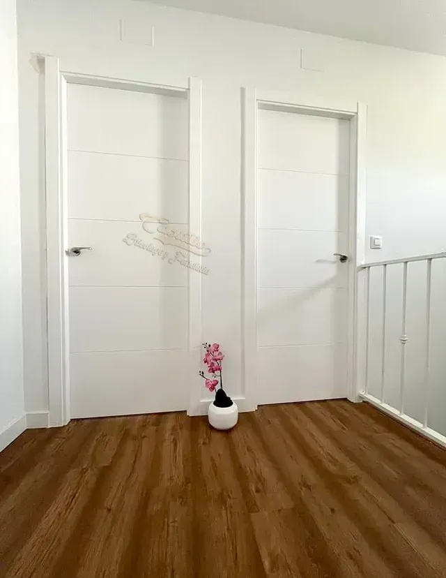 Un pasillo con dos puertas blancas y un jarrón con flores en el suelo.