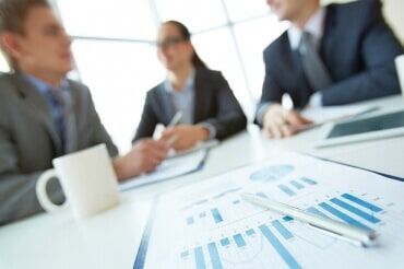 Reunião de negócios com gráficos, uma caneta e três pessoas de terno desfocadas ao fundo.