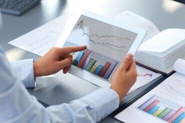 Pessoa usando um tablet exibindo gráficos e tabelas financeiras; cercada por documentos.