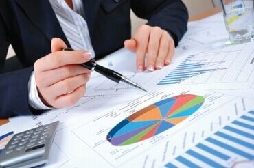 Uma pessoa de terno analisa gráficos e tabelas financeiras, apontando com uma caneta.