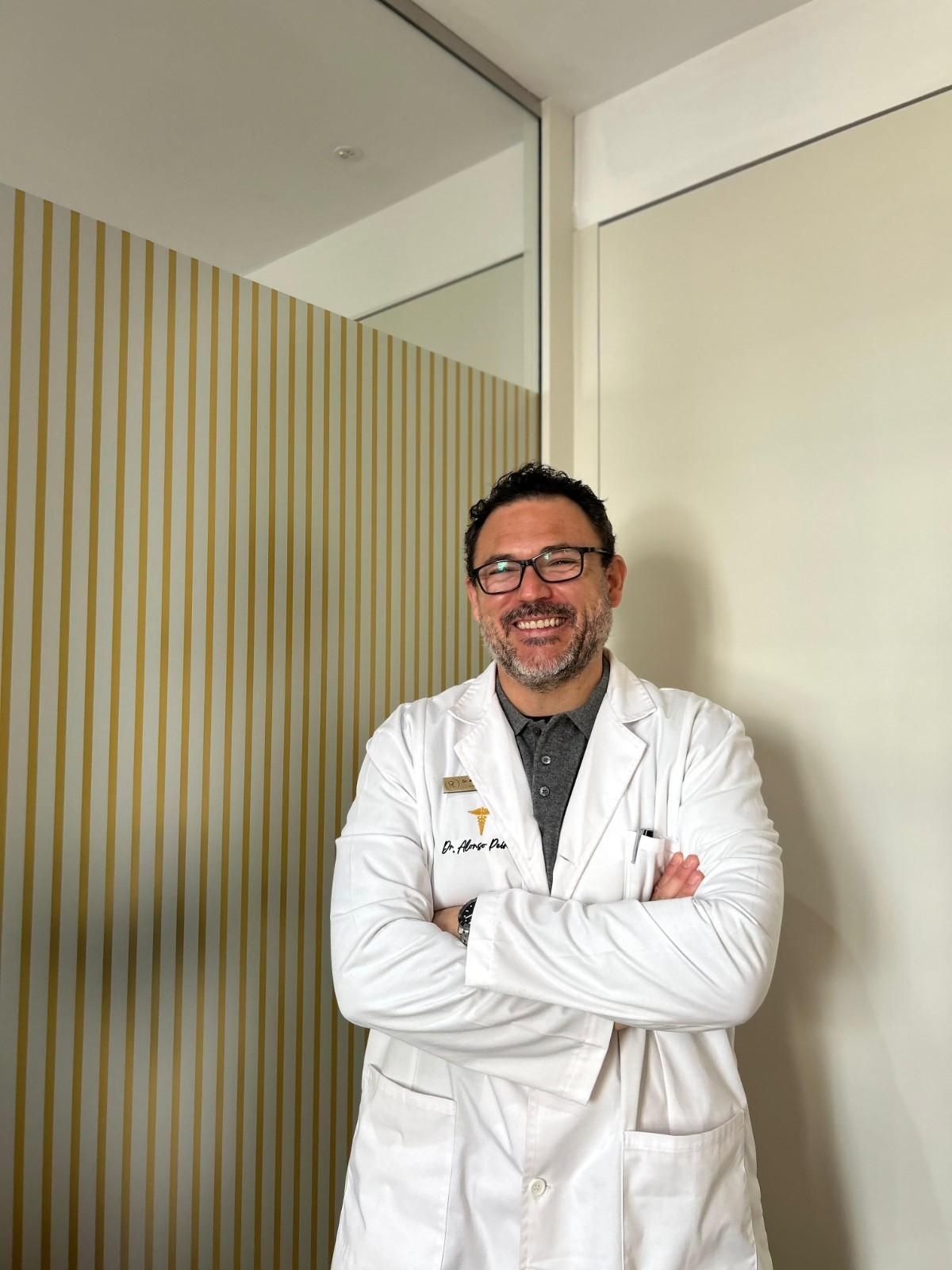 Hombre con bata blanca, brazos cruzados y sonriente. Apoyado en una pared revestida de madera.