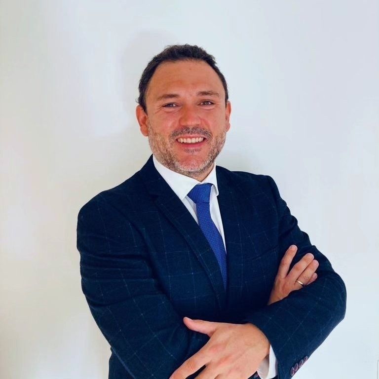 Hombre con traje azul marino y corbata azul, con los brazos cruzados, sonriendo, sobre un fondo blanco.