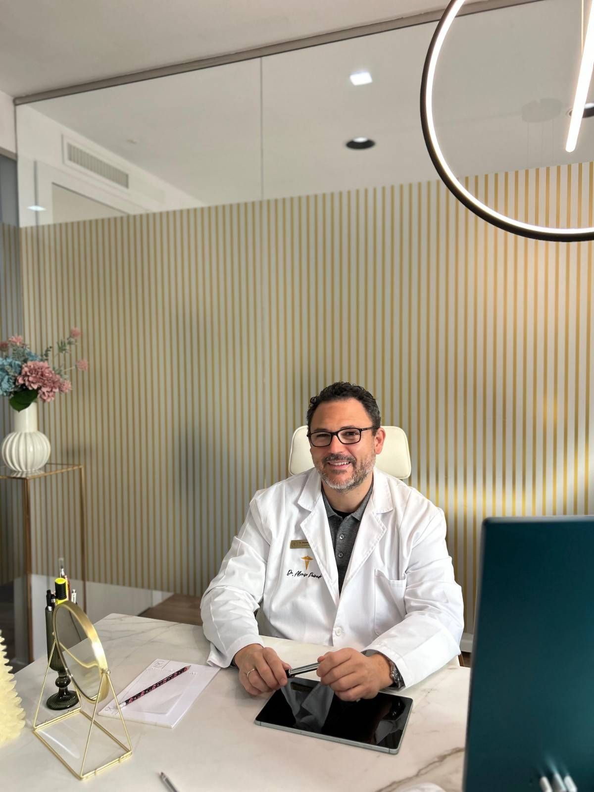 Hombre con bata blanca sentado en un escritorio en la oficina, sonriendo, con objetos en la mano. 