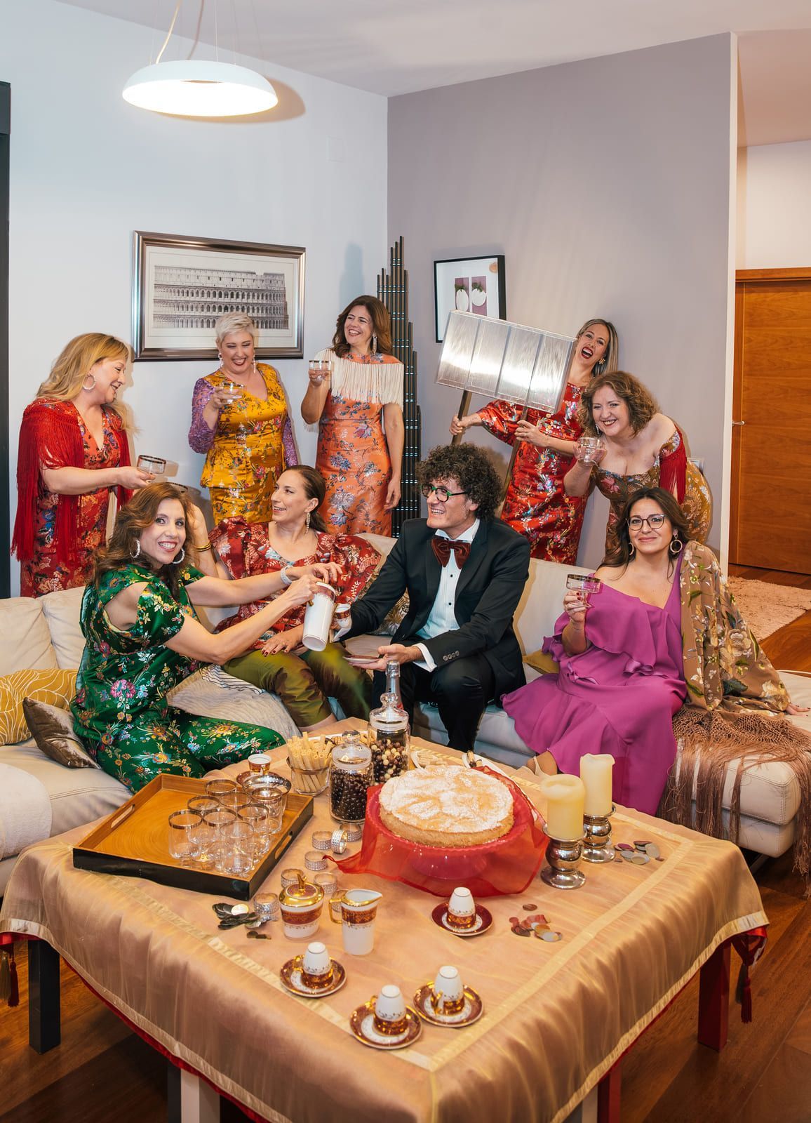 Grupo de personas en una sala de estar, sonriendo, bebiendo y con bebidas en la mano. Una mesa con comida y bebidas frente a ellos.