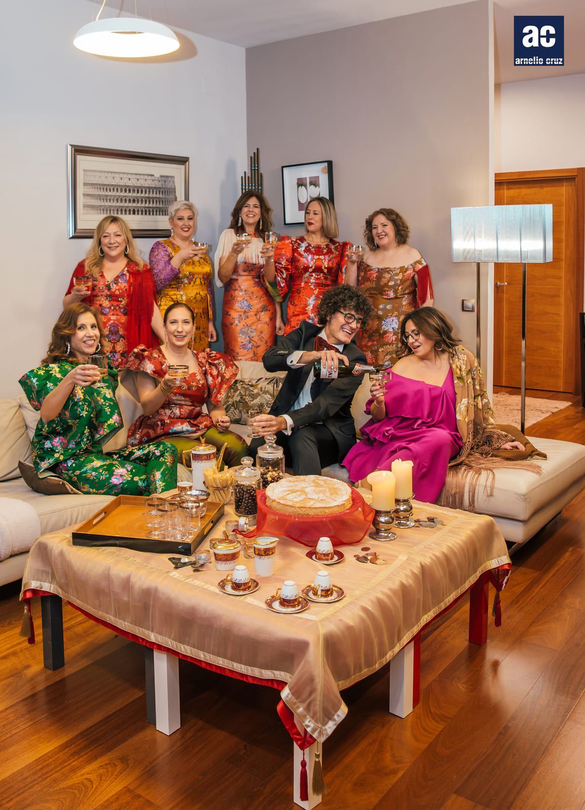 Grupo de personas con vestidos vibrantes socializando alrededor de una mesa con café y golosinas en una sala de estar.