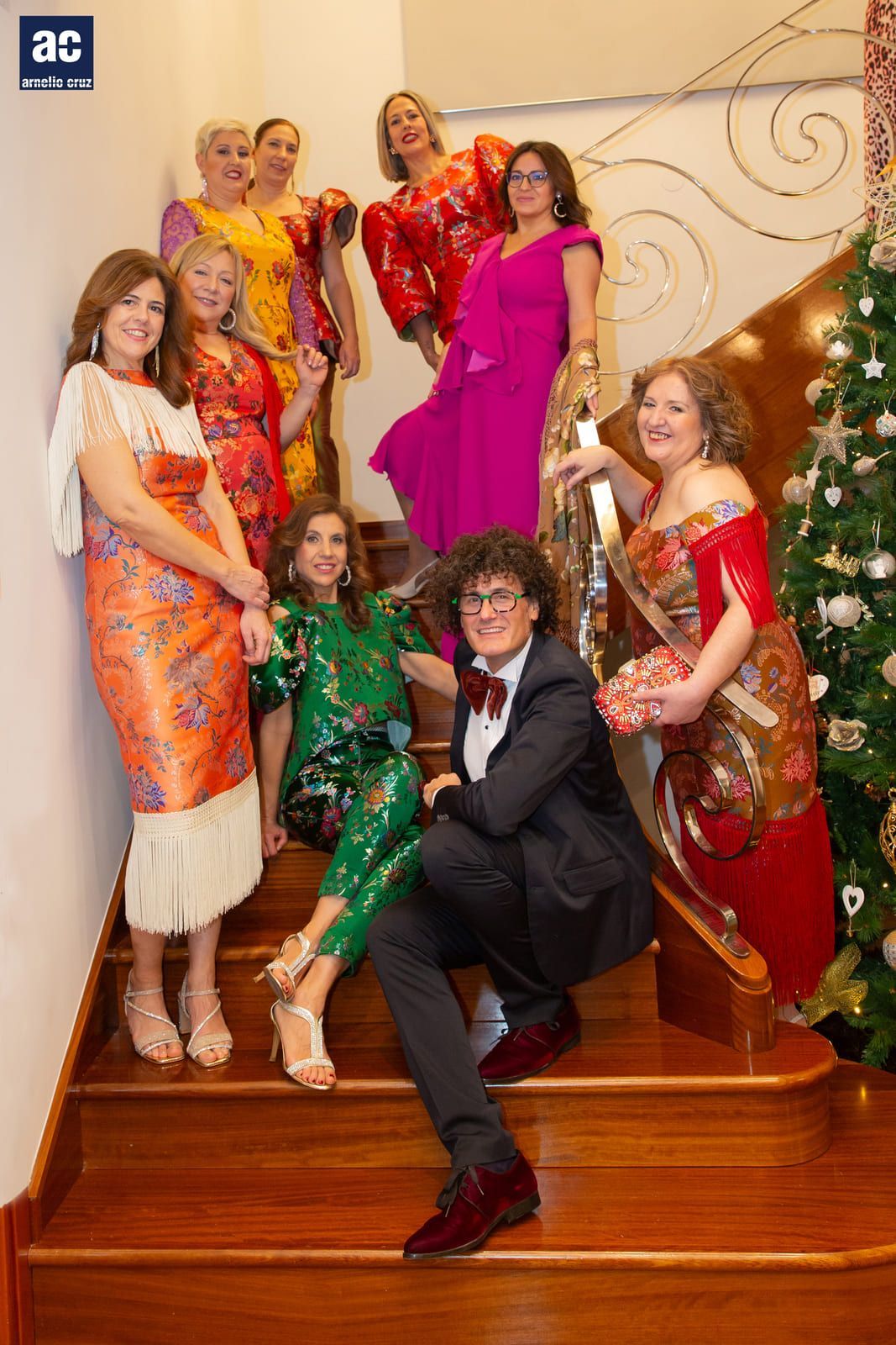 Grupo de personas posando en una escalera, con atuendo festivo. Un hombre de pelo rizado y pajarita está sentado en primer plano.