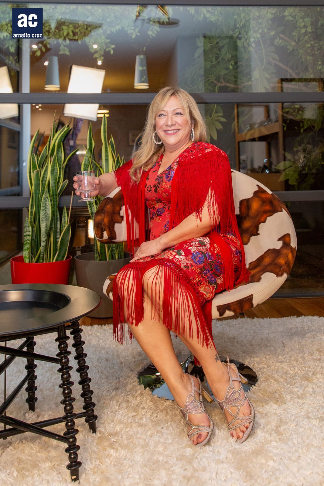 Mujer con vestido rojo y chal, sosteniendo un vaso, sentada en una silla estampada, sonriendo.