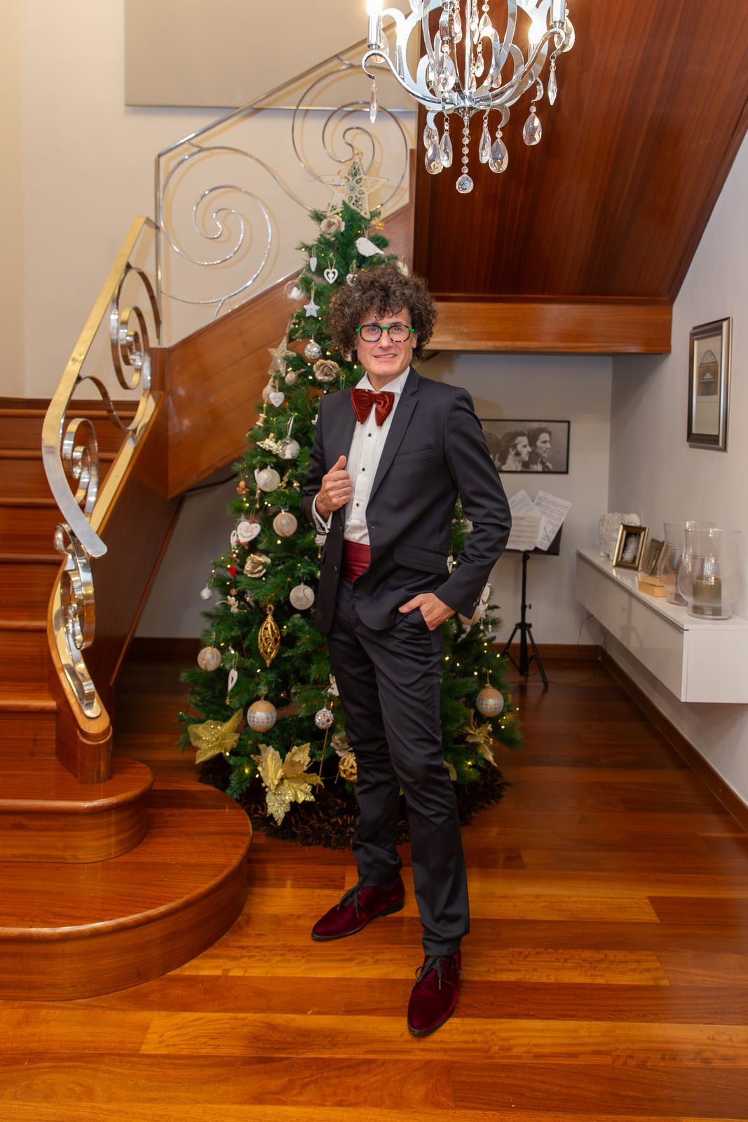 Persona con traje y pajarita, posando junto a un árbol de Navidad, con una escalera y una lámpara de araña al fondo.