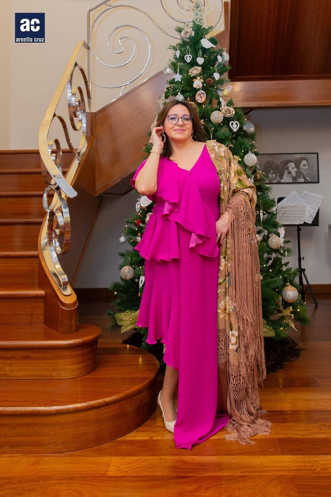 Una mujer con un vestido y un chal rosa se encuentra junto a un árbol de Navidad decorado y una escalera de madera.