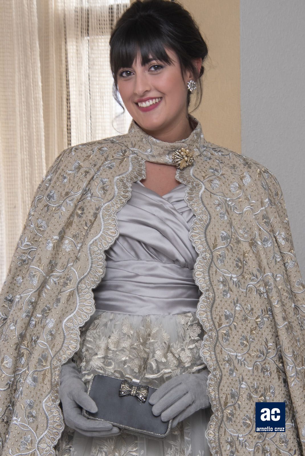 Mujer con vestido plateado y capa ornamentada, sonriendo, sosteniendo un bolso de mano, con guantes; ambientación interior.
