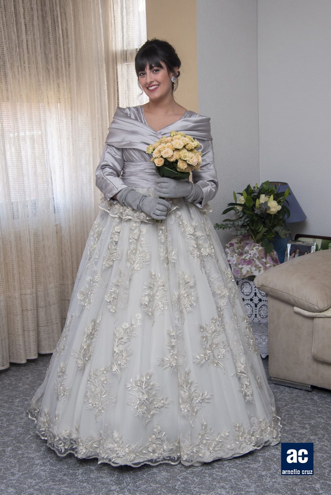 Mujer con vestido de novia plateado sosteniendo un ramo, de pie en el interior cerca de una ventana.