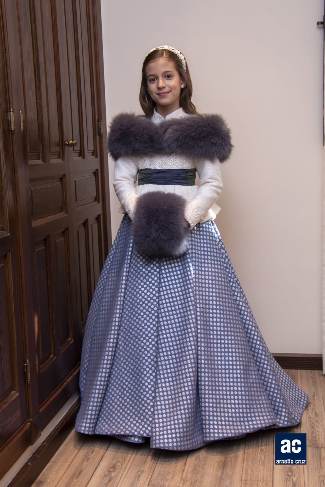 Persona joven con un vestido formal, estola de piel y manguito, de pie en el interior.