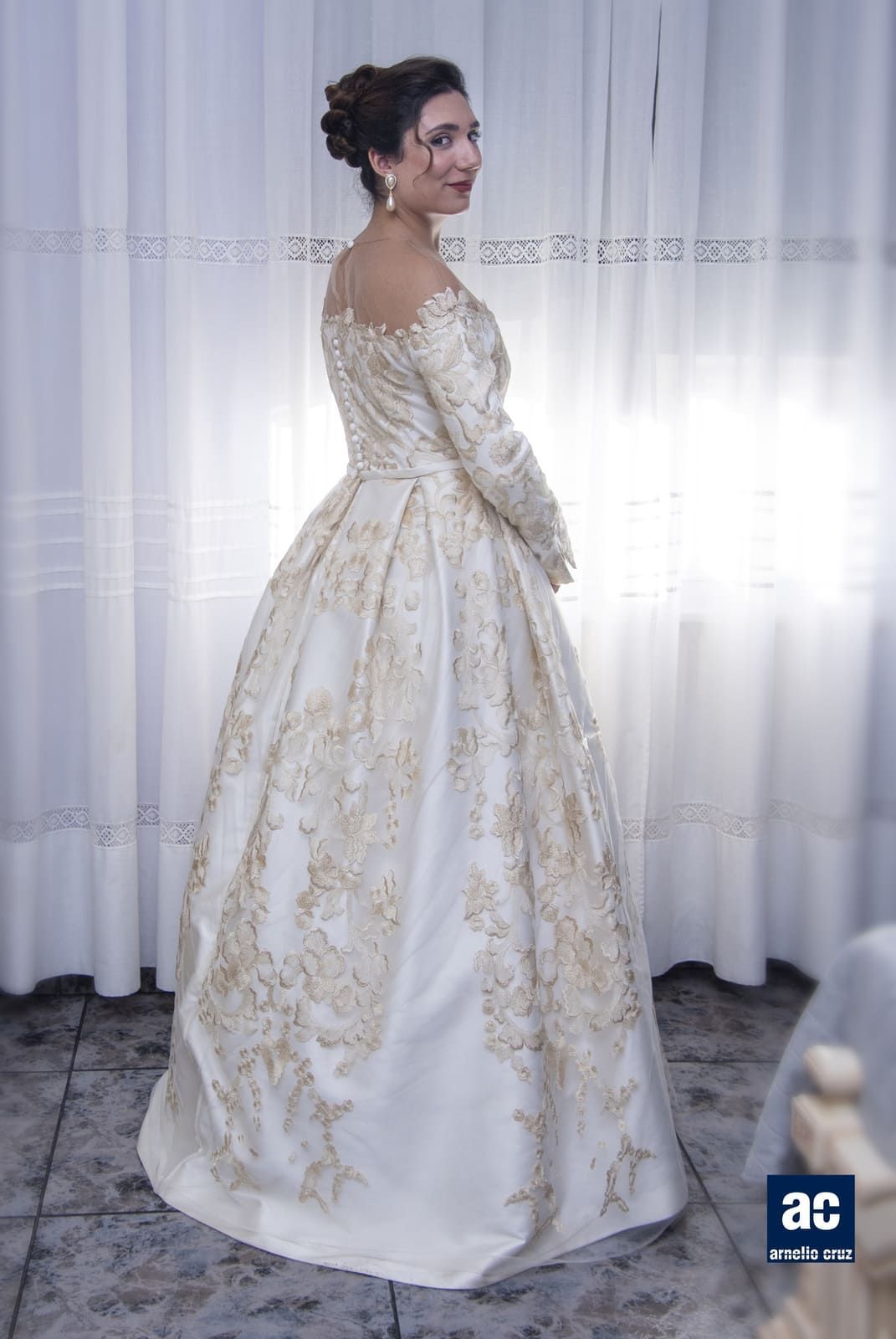 Una mujer con un vestido de fiesta blanco y dorado sonríe, de pie junto a una cortina transparente.