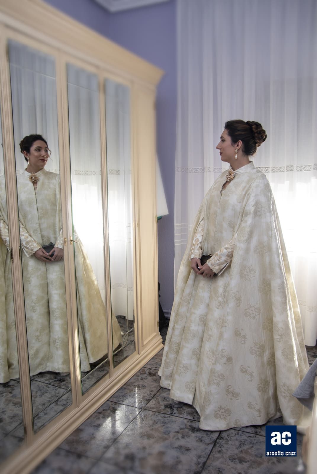 Mujer con vestido y capa color crema adornados, mirándose en el espejo; habitación iluminada.