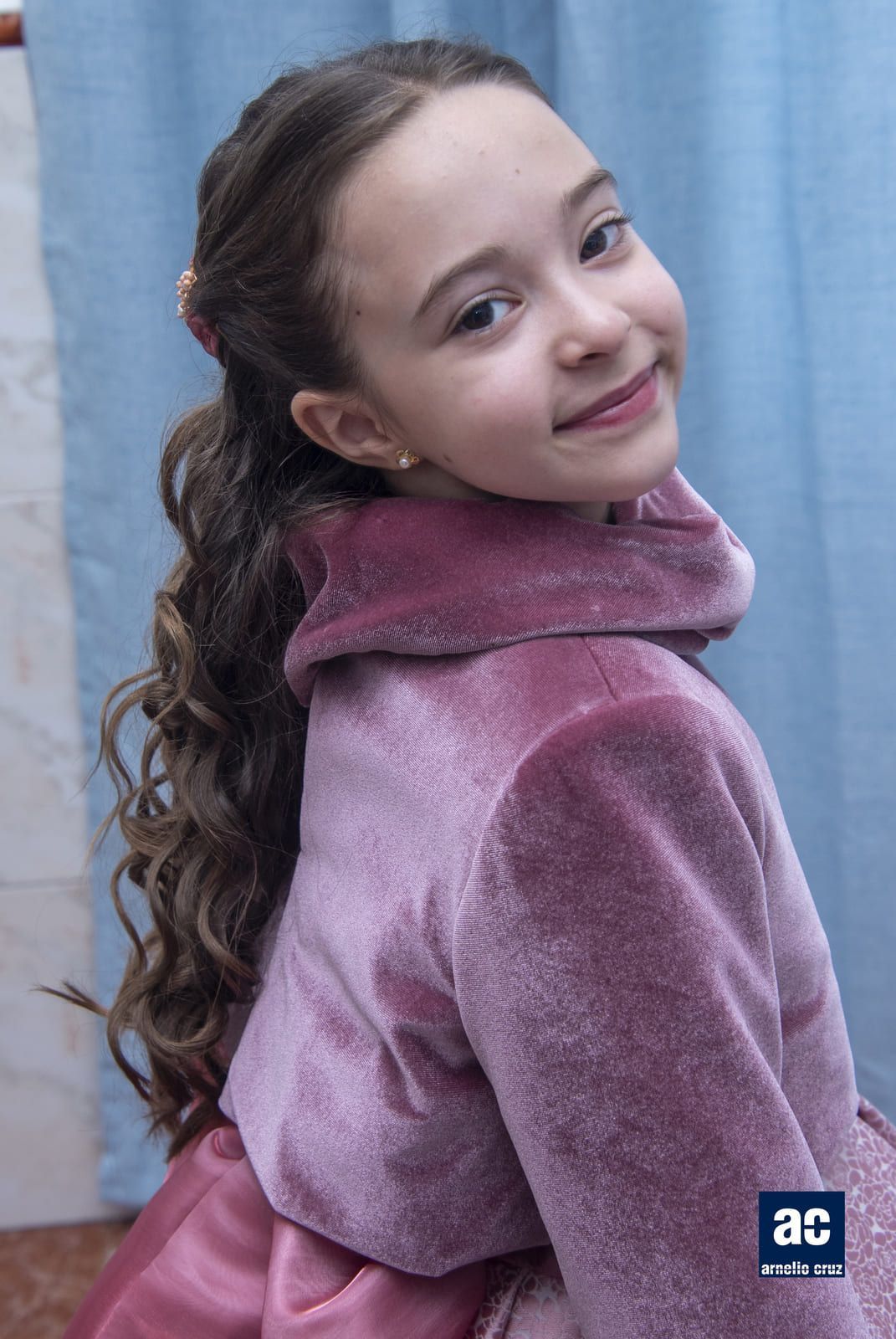 Chica joven con abrigo y vestido de terciopelo rosa, sonriendo. Lleva el pelo castaño y rizado recogido a media pierna.