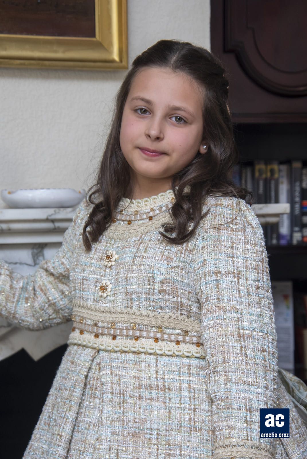 Chica joven con abrigo de tweed, de pie junto a una chimenea, sonriendo levemente.