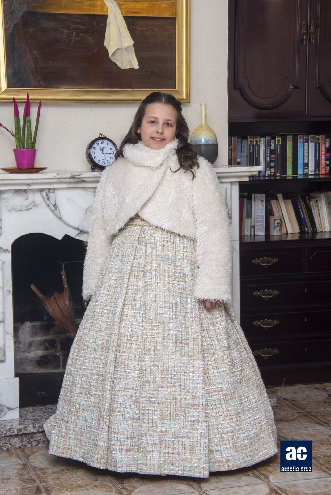 Una persona joven con un vestido blanco formal y una chaqueta se encuentra frente a una chimenea.