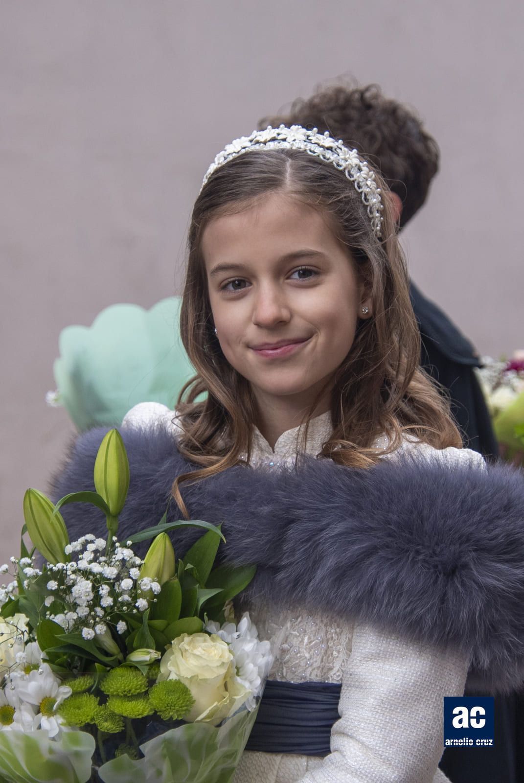Persona joven sosteniendo flores, vistiendo una estola de piel y una diadema adornada, sonriendo.