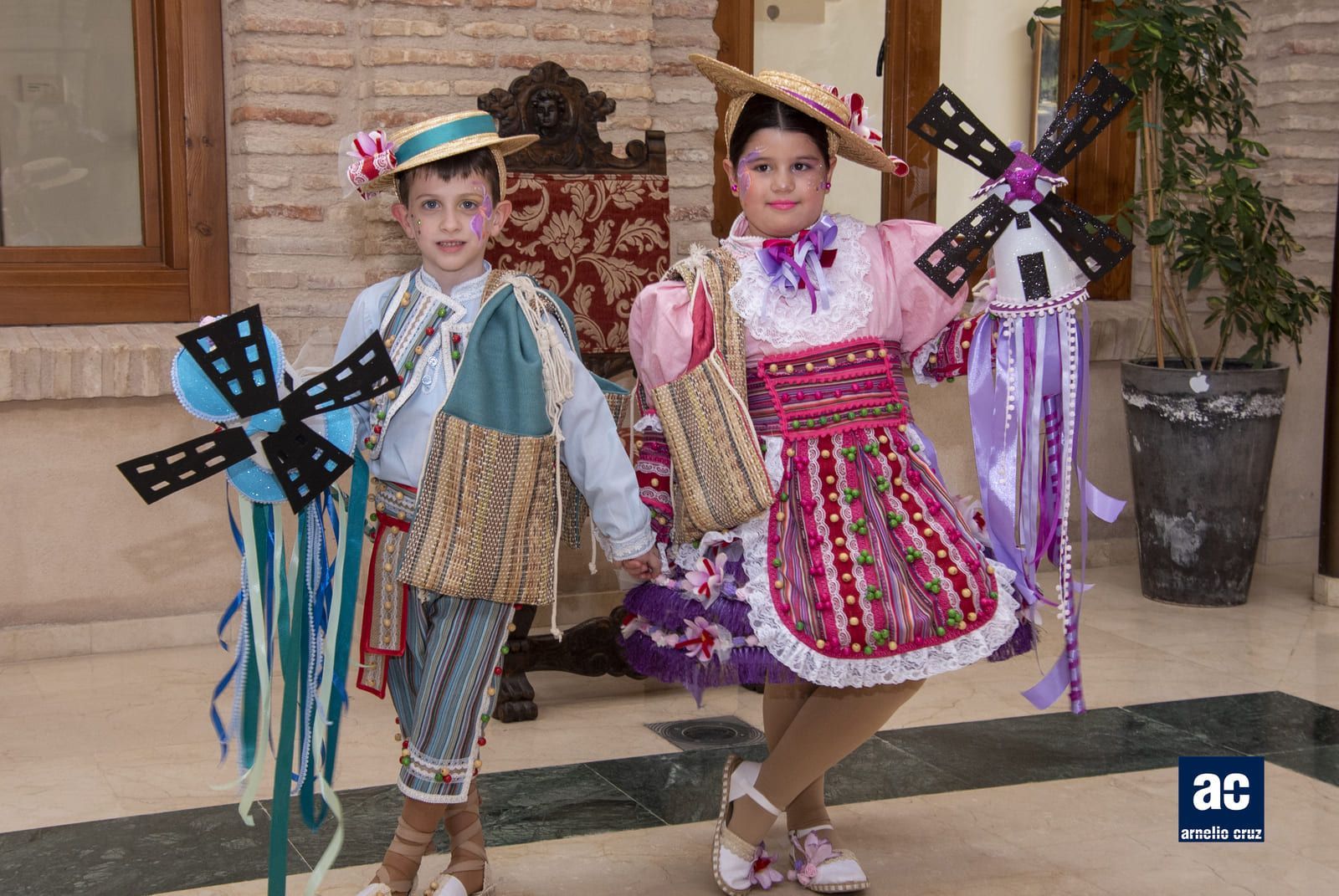 Dos niños con trajes tradicionales sostienen molinos de viento de juguete. Están dentro, cerca de una ventana.