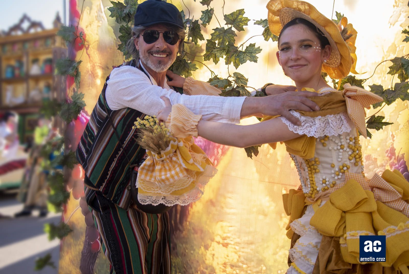Hombre a rayas y mujer de amarillo, bailando al aire libre, sonriendo. Luz del sol y follaje.