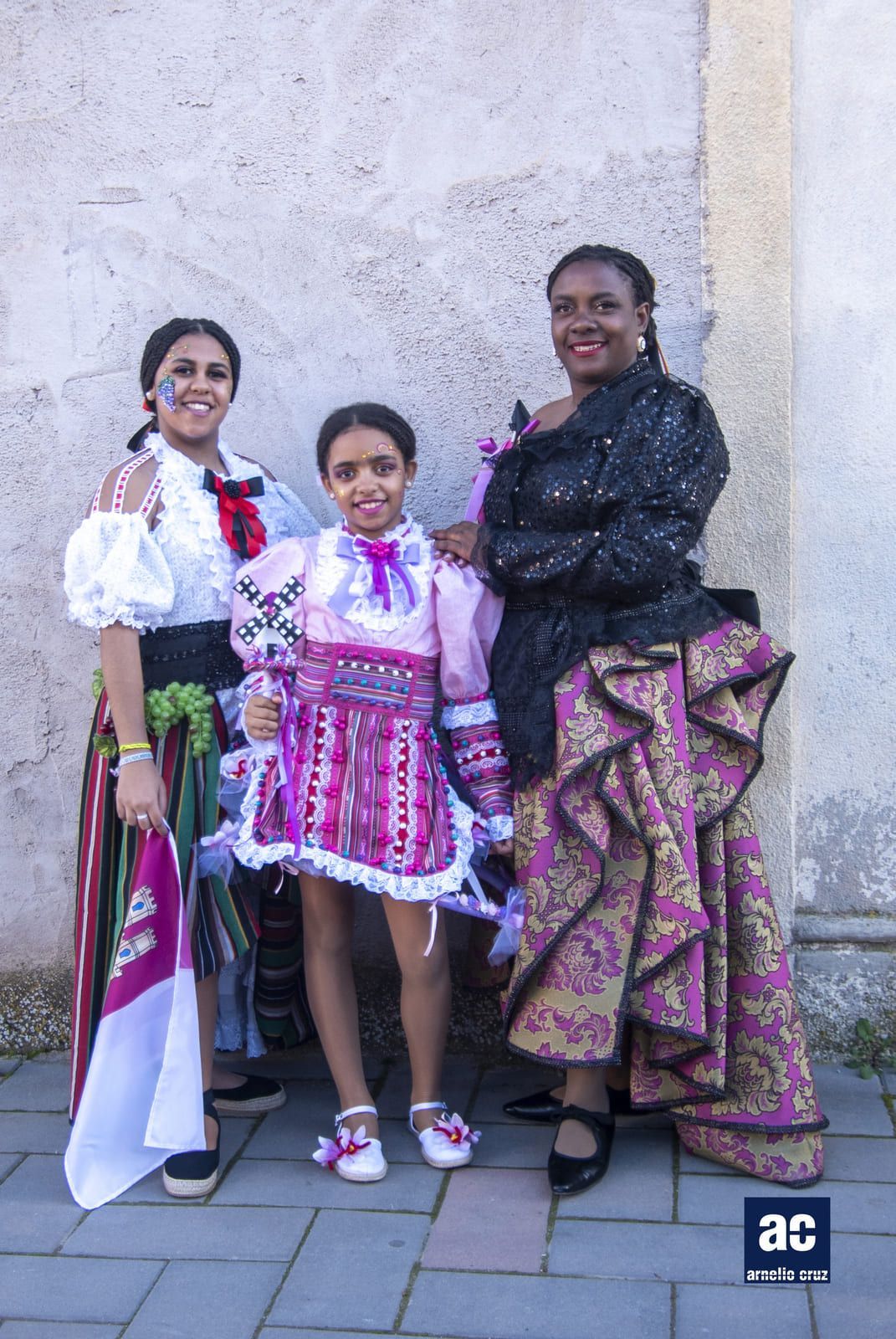 Tres personas con atuendos festivos posando al aire libre. El niño del medio lleva un vestido rosa, y los adultos lucen trajes elaborados y estampados.