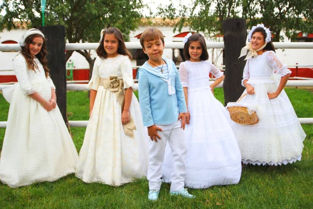 Un niño con una camisa azul se encuentra entre dos niñas con vestidos blancos.